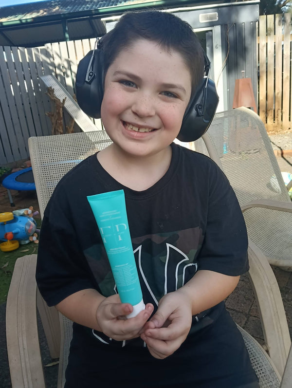 Smiling boy wearing earmuffs in the backyard, holding a tube of Flossy and Pure toothpaste